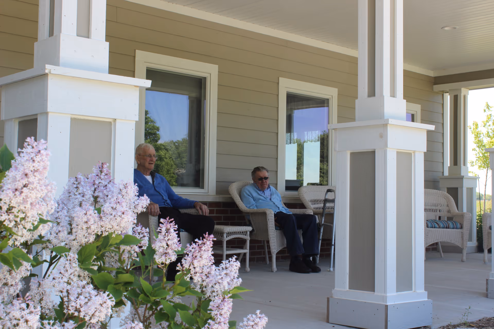 Two elderly men sitting on wicker chairs on a covered porch of a senior living facility, with blooming lilac bushes in the foreground and beige siding on the building.