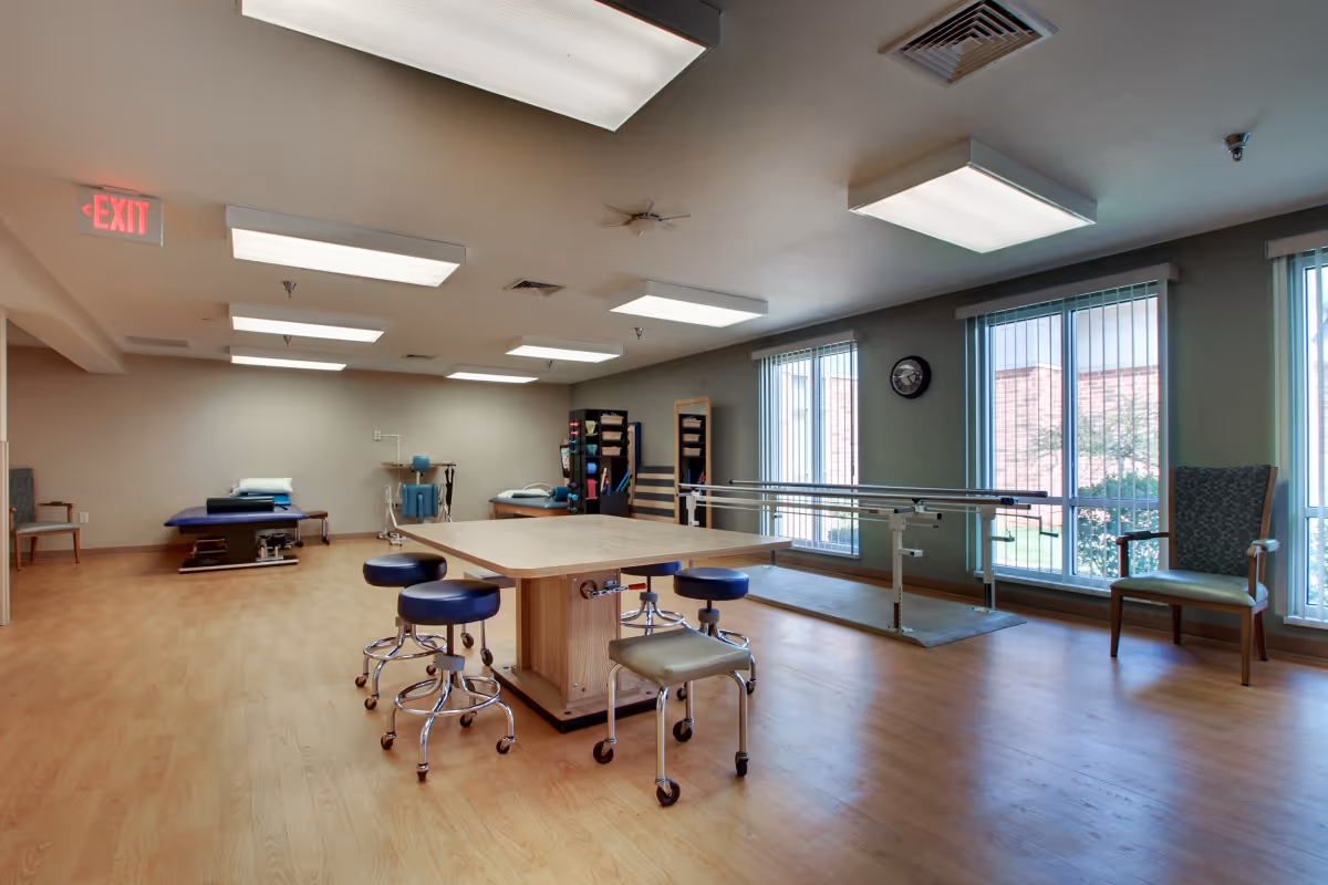 A spacious physical therapy room with wooden flooring, a large central table surrounded by stools on wheels, parallel bars for walking exercises near large windows with vertical blinds, therapy beds, exercise equipment, and chairs along the walls. The room is well-lit with ceiling lights and has an exit sign visible.
