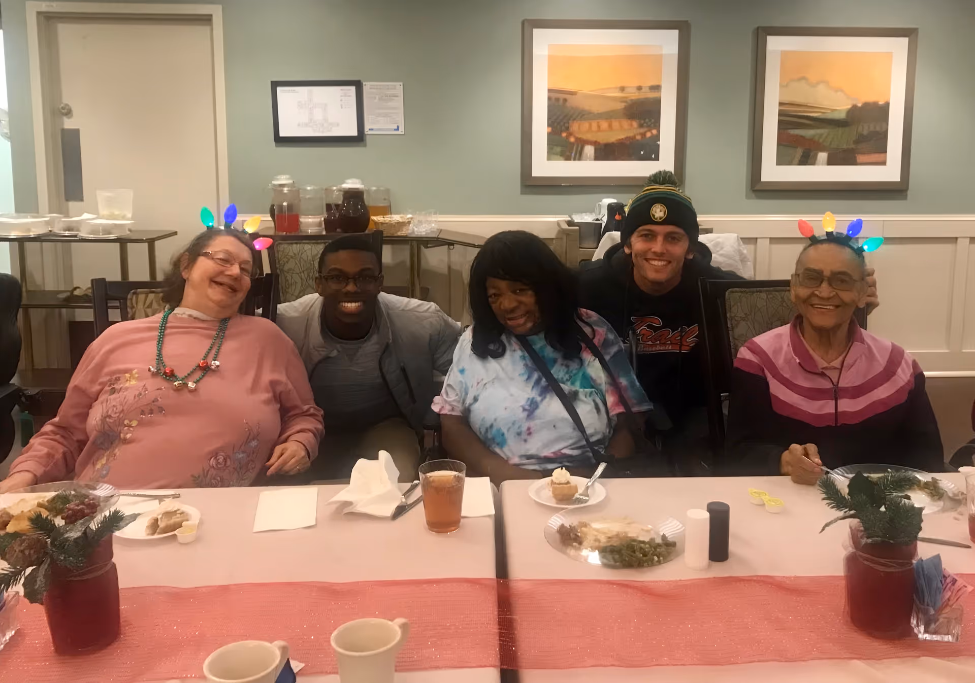 Five people smiling and seated at a decorated dining table with plates of food, holiday light headbands, and framed artwork on the wall behind them.