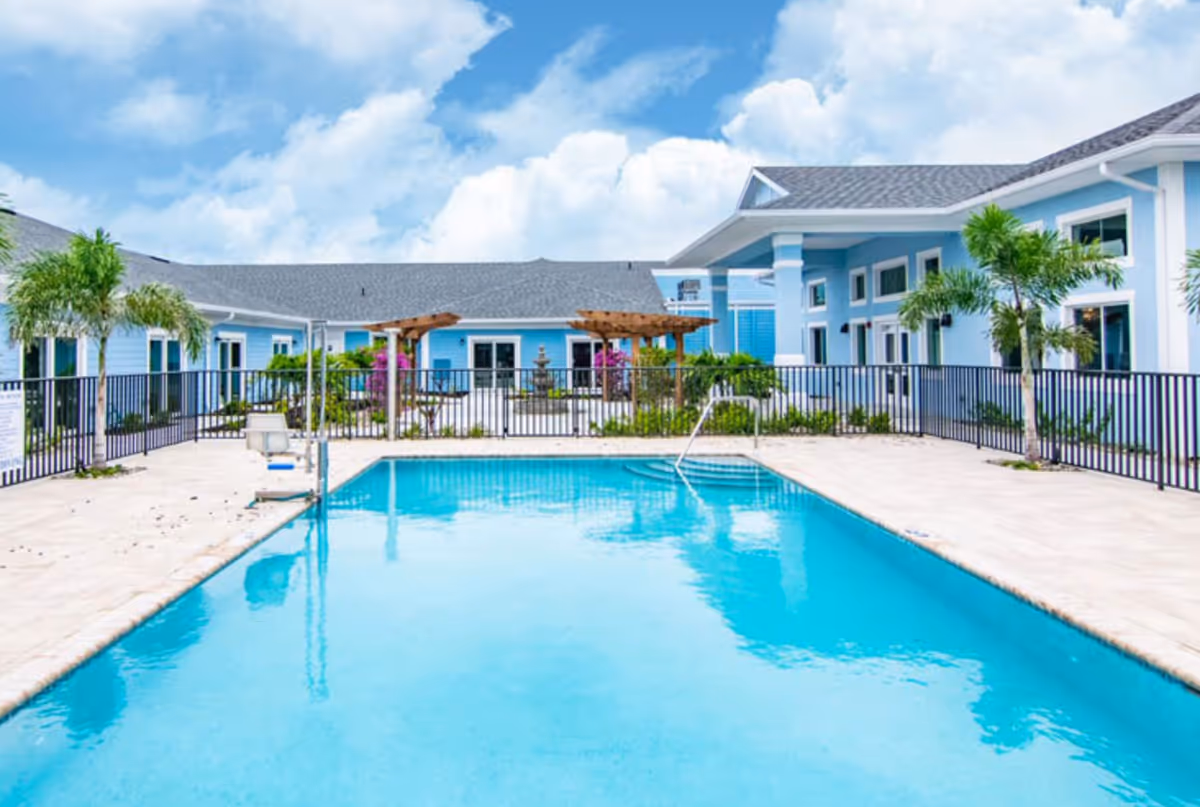 Outdoor swimming pool with clear blue water surrounded by a light-colored tiled deck and black metal fencing. The pool area is part of a light blue building complex with white trim, palm trees, and a pergola with greenery and flowers. The sky is partly cloudy with blue patches.