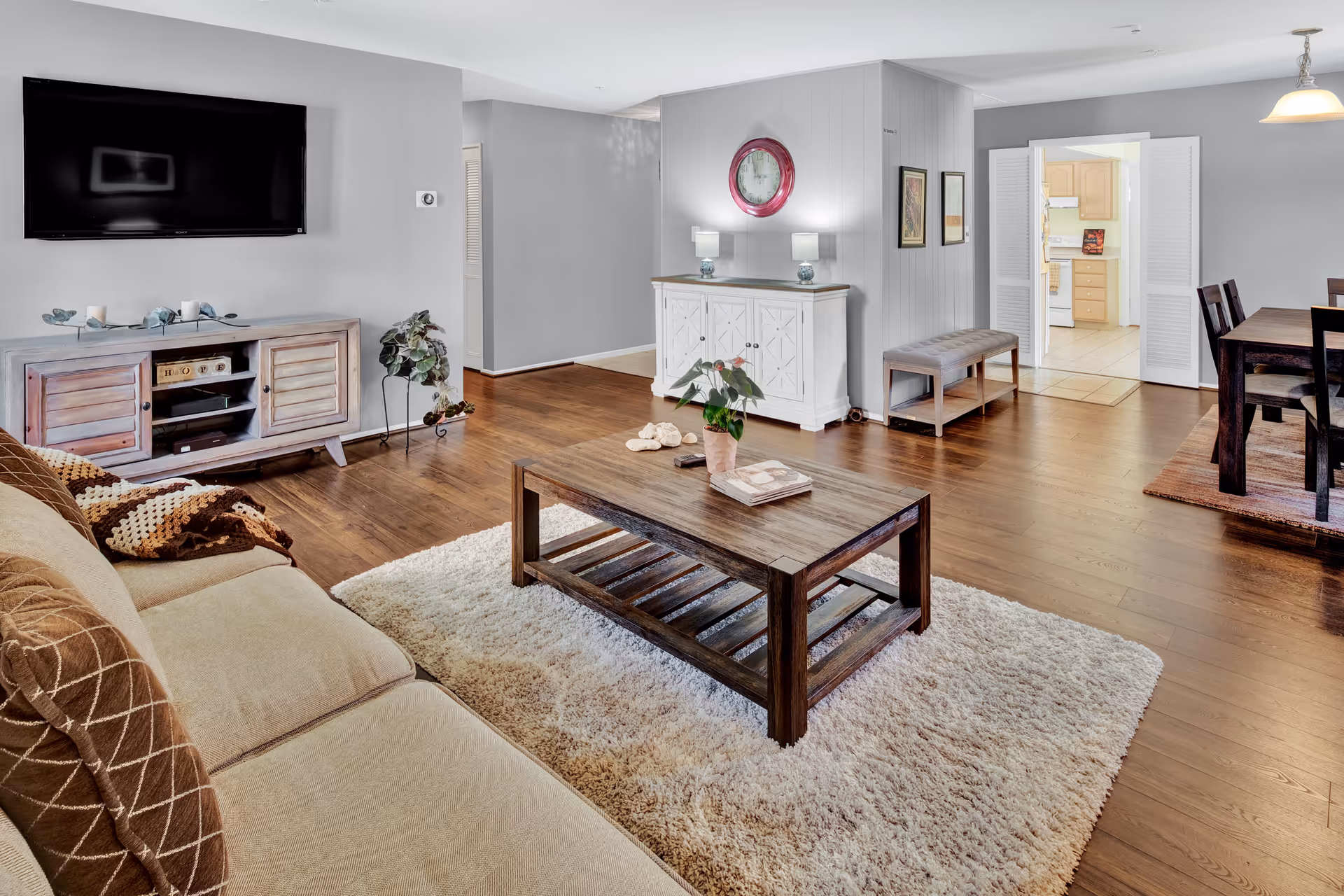 A spacious living room with a beige sofa adorned with brown and beige pillows and a knitted throw. A wooden coffee table sits on a light-colored shag rug in the center. Against the wall is a wooden media console with a flat-screen TV mounted above it. The room has wooden flooring and light gray walls. In the background, there is a white cabinet with two lamps and a large round clock above it, a bench, and a dining area with a dark wooden table and chairs. A doorway leads to a kitchen with light wood cabinets.