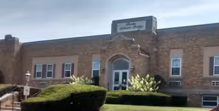 Exterior front view of a brick building with a sign above the entrance that reads 'Berk's Leisure Living'. The building has several windows and is surrounded by neatly trimmed bushes and greenery under a clear sky.