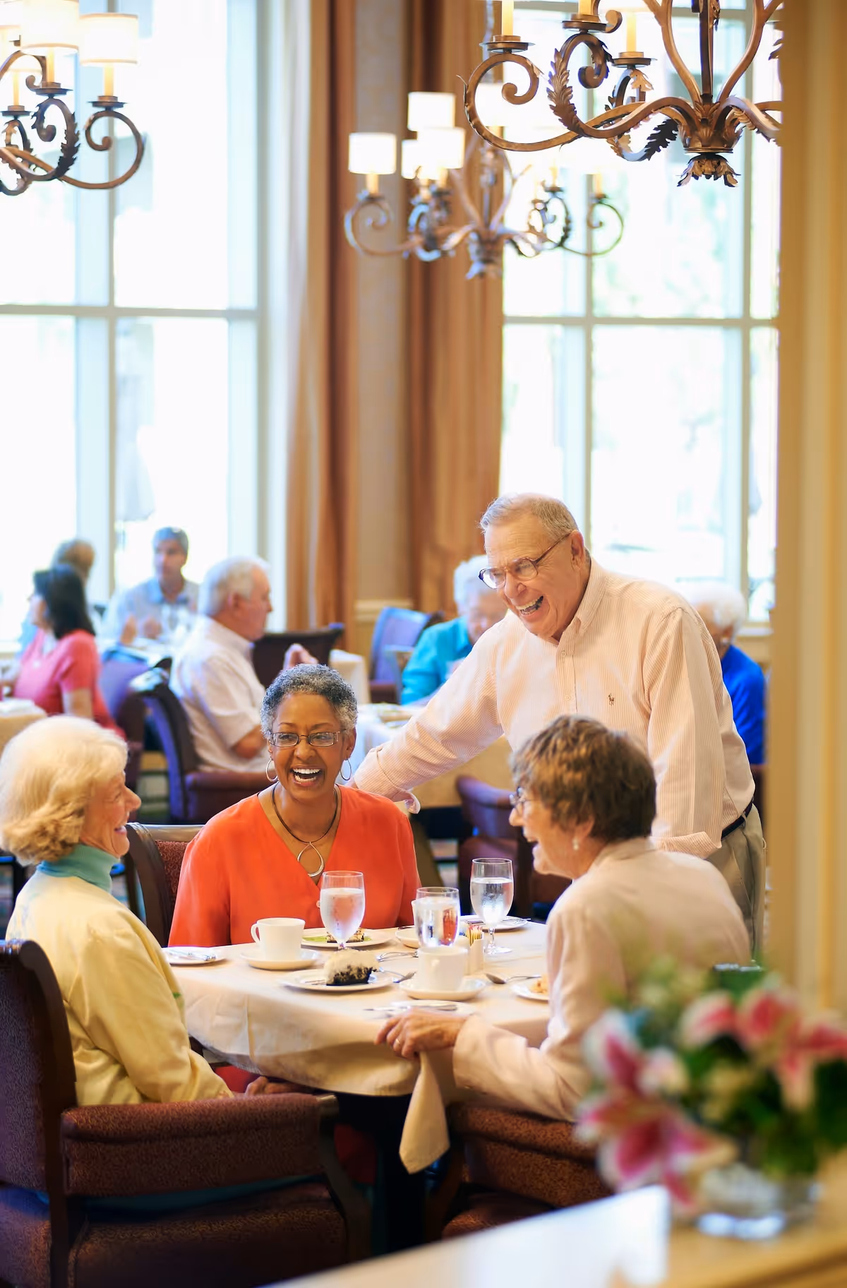 A group of elderly people enjoying a meal and conversation together in a bright dining room with large windows and chandeliers.