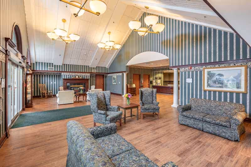 A spacious and well-lit common area with high vaulted ceilings and striped wallpaper. The room features multiple upholstered sofas and armchairs arranged around small tables, a fireplace in the background, and large windows and glass doors on the left side letting in natural light. There is a green carpet near the entrance and wooden flooring throughout the space.