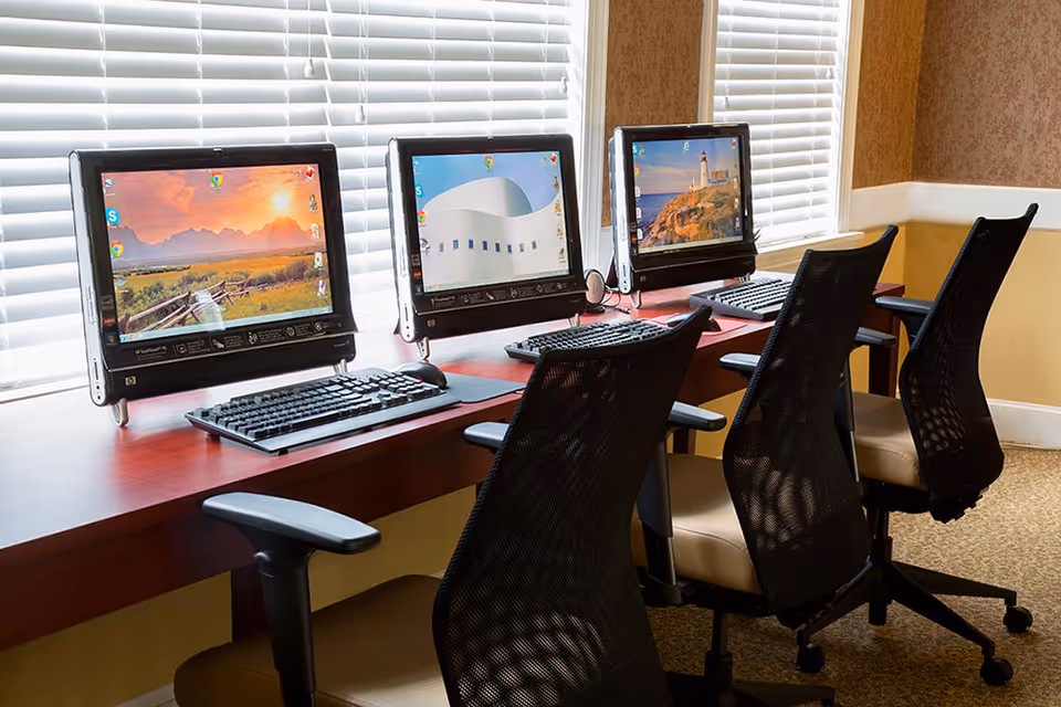 A row of three desktop computers on a wooden desk with black mesh office chairs in front of each. The computers display different scenic wallpapers and are positioned in front of windows with white blinds. The room has beige walls with white trim and carpeted flooring.
