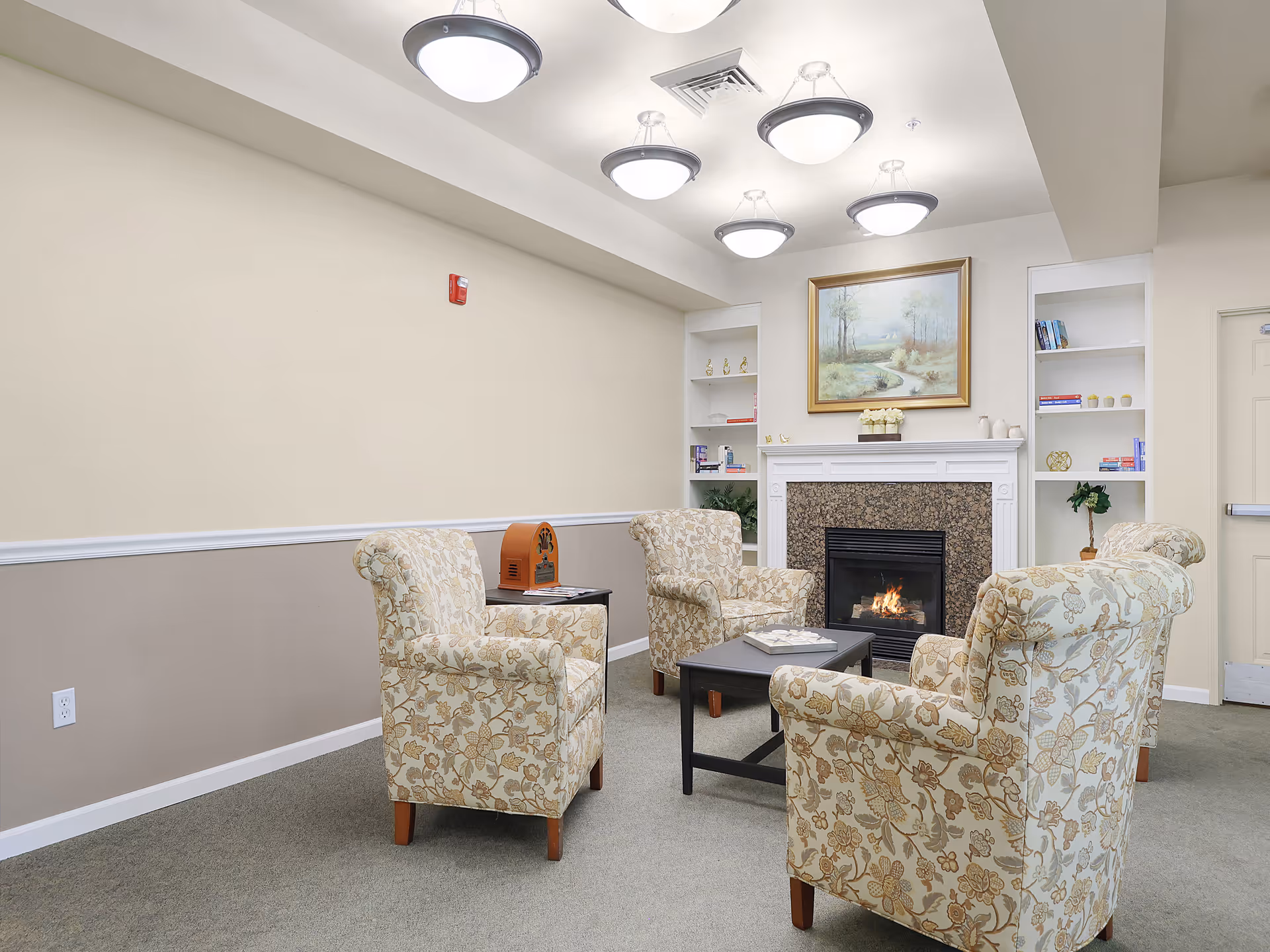 A cozy sitting area with four floral-patterned armchairs arranged around a black coffee table in front of a lit fireplace. The fireplace has a white mantel with decorative items and built-in shelves on either side holding books and small plants. Above the fireplace hangs a framed painting of a serene landscape. The room has soft beige walls, a carpeted floor, and ceiling lights.