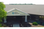 Entrance to Adams Woodcrest nursing facility showing a covered drop-off area with a sign that reads 'Adams Woodcrest Nursing Entrance'. The building has a sloped roof and is surrounded by some greenery and bushes.