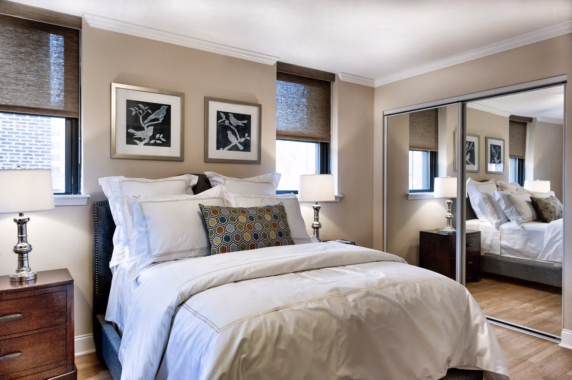 A neatly made bed with white linens and multiple pillows, including a decorative pillow with a hexagonal pattern, is positioned between two windows with brown blinds. There are two framed bird artworks on the beige wall above the bed. On either side of the bed are wooden nightstands with silver lamps. A large mirrored sliding closet door reflects the room.