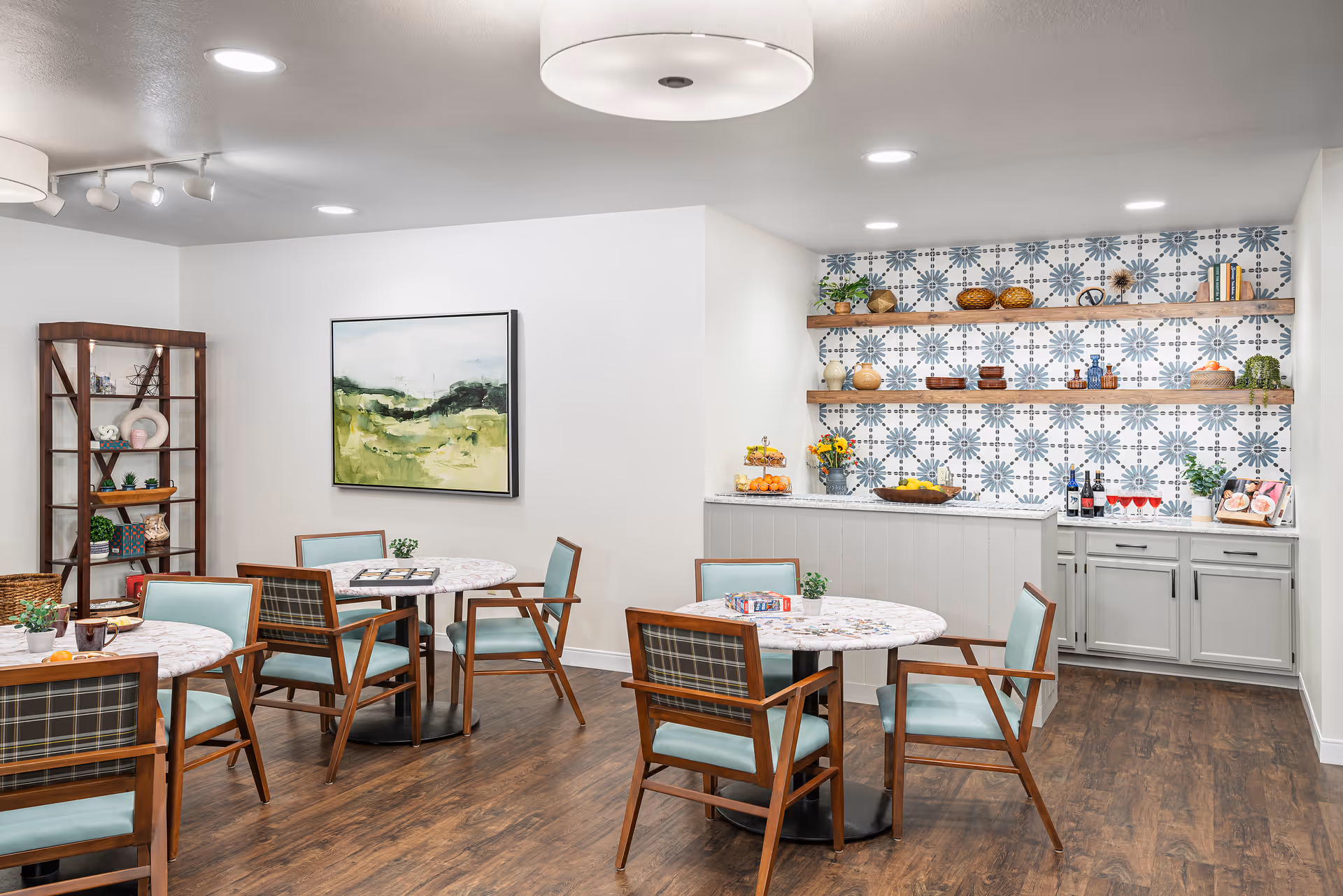 A cozy dining area with round marble tables and wooden chairs with light blue cushions. The room features a wooden floor, a decorative shelving unit with various items, and a counter with open wooden shelves above it displaying decorative bowls, plants, and books against a patterned blue and white tile wall. The space is well-lit with ceiling lights and a large circular ceiling fixture.