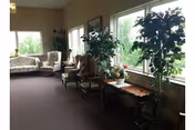 A cozy interior common area with large windows letting in natural light. The room features several upholstered chairs and small wooden tables, along with tall potted plants placed near the windows. The walls have a patterned wallpaper and the floor is carpeted in a dark color.