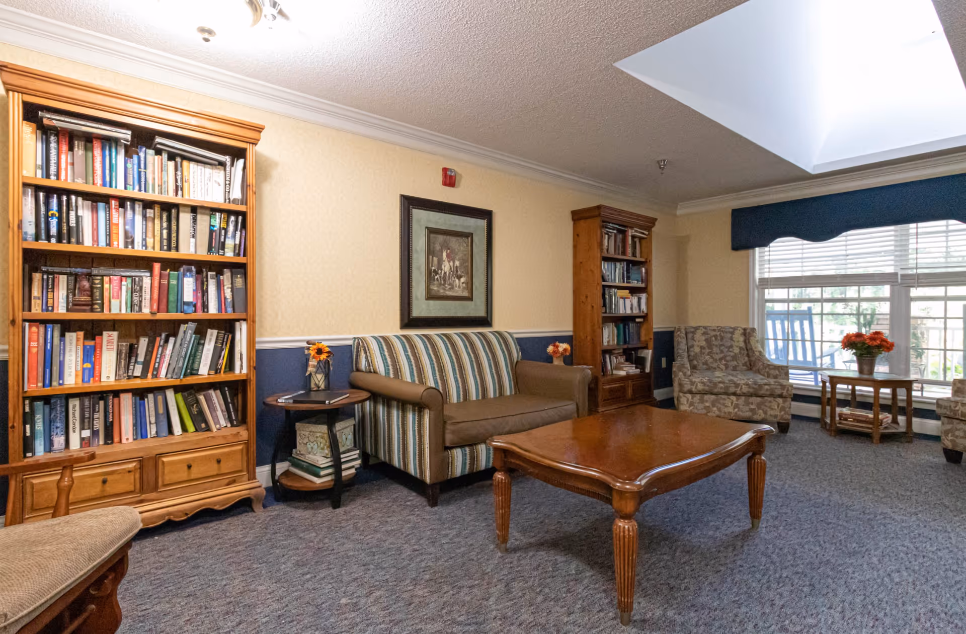 A cozy living room area with a striped loveseat, two floral armchairs, a wooden coffee table, two wooden bookshelves filled with books, a small round side table with a flower vase, a large window with blinds and a blue valance, and a skylight in the ceiling.