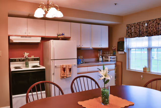 Small kitchen with white cabinets, refrigerator and stove, and a dining table in the foreground with a vase of white lilies.