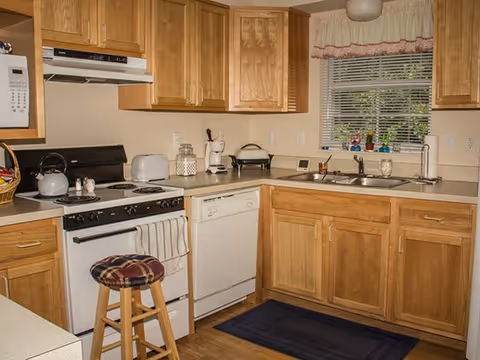 Small kitchen with wooden cabinets, white appliances, a stool, and a window above the sink.