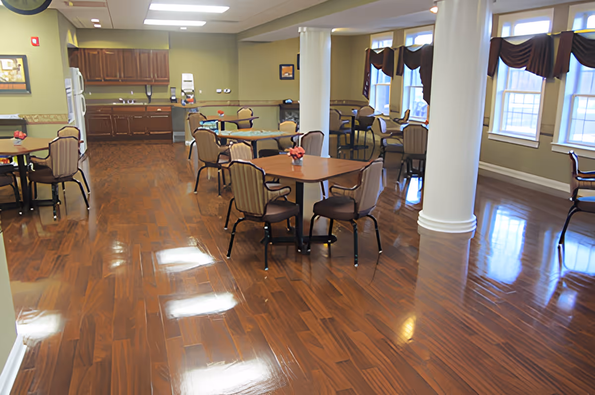 A spacious dining area with multiple tables and chairs arranged on a polished wooden floor. The room features large white columns, windows with dark valances, and a kitchen area with wooden cabinets and a refrigerator in the background.
