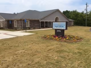 Exterior view of a single-story brick building with a gray roof, surrounded by a grassy lawn. In front of the building, there is a sign that reads 'Angels Divine Personal Care Home' with the number 3065 below it, and a flower bed with colorful flowers around the sign.