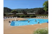 Outdoor swimming pool with several people swimming and relaxing in the water, surrounded by lounge chairs and greenery with hills in the background under a clear blue sky.