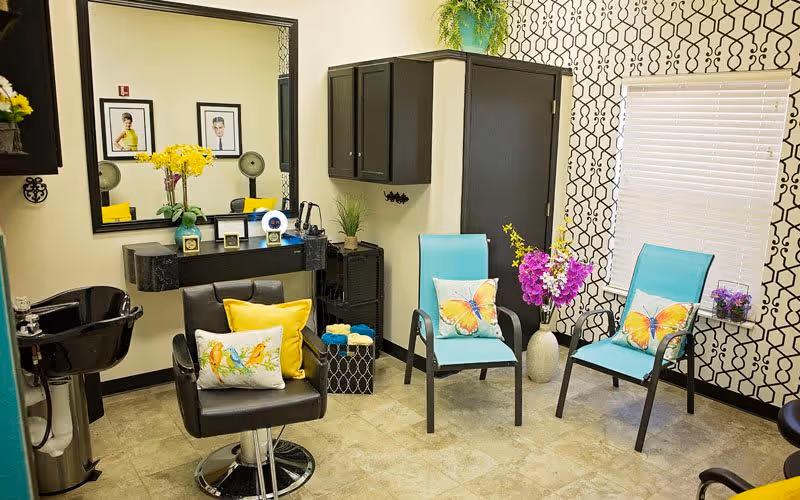A brightly lit salon room with a black styling chair adorned with colorful pillows, a black sink for washing hair, a large mirror above a small black counter with yellow flowers and framed photos, two turquoise chairs with butterfly pillows, a small side table with purple flowers, and patterned black and white wallpaper on one wall.