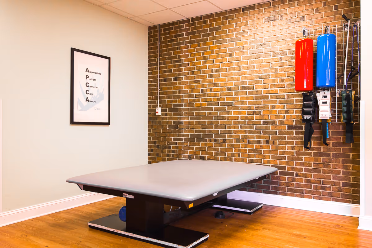 A therapy or exercise room with a padded treatment table in the center, a brick wall in the background, and various exercise equipment hanging on the wall including red and blue cylindrical pads and resistance bands. A framed poster with the acronym APCCA is mounted on the adjacent white wall.