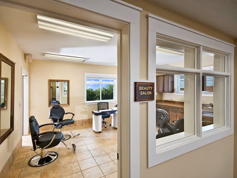Interior view of a beauty salon in a senior living facility with two black salon chairs, a desk with a chair, mirrors on the walls, and large windows letting in natural light.