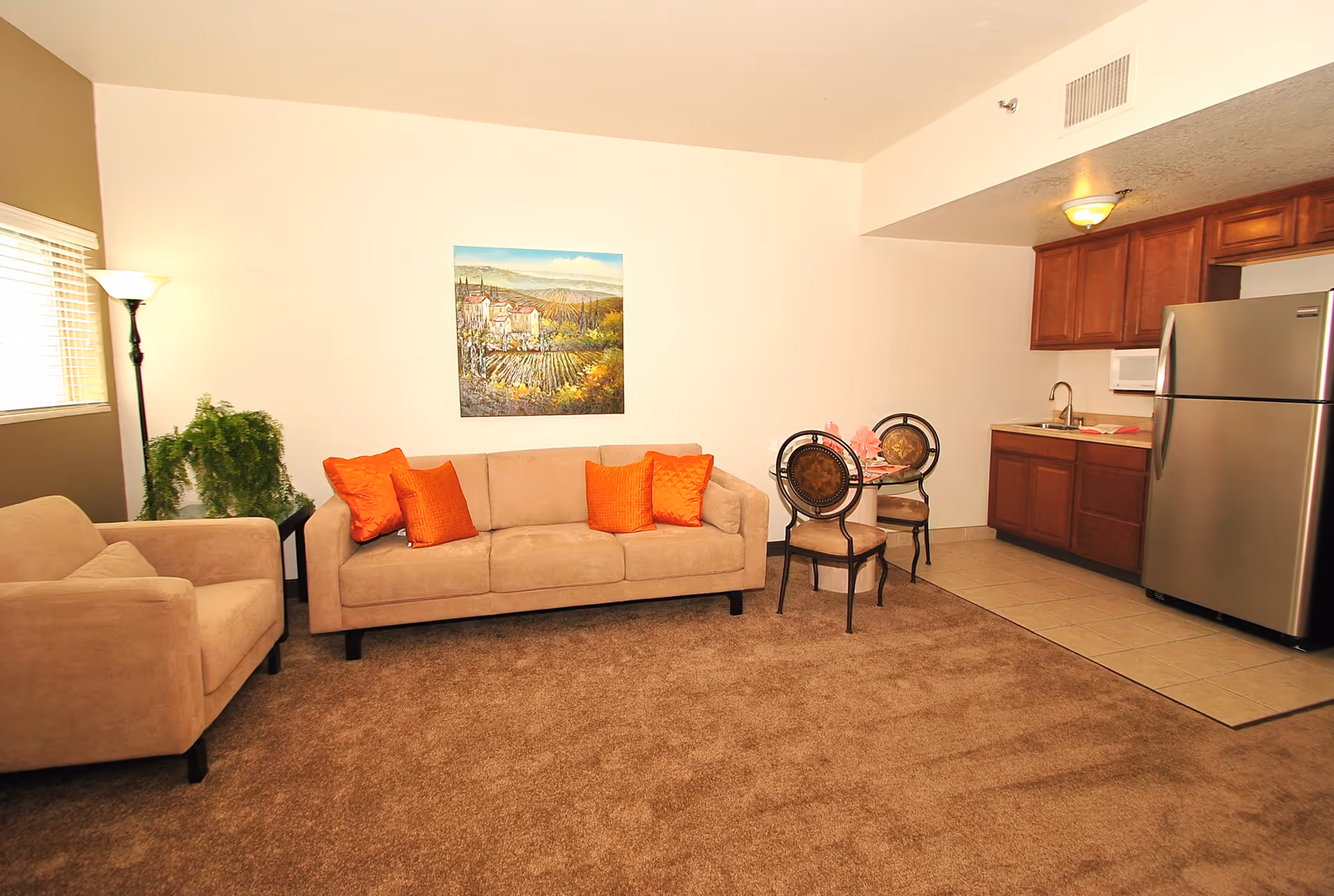 A cozy living area with a beige sofa adorned with orange pillows, a matching armchair, a small round dining table with two chairs, and a compact kitchen featuring wooden cabinets, a stainless steel refrigerator, and a microwave. A floor lamp and a green plant are near the window, and a landscape painting hangs on the wall above the sofa.