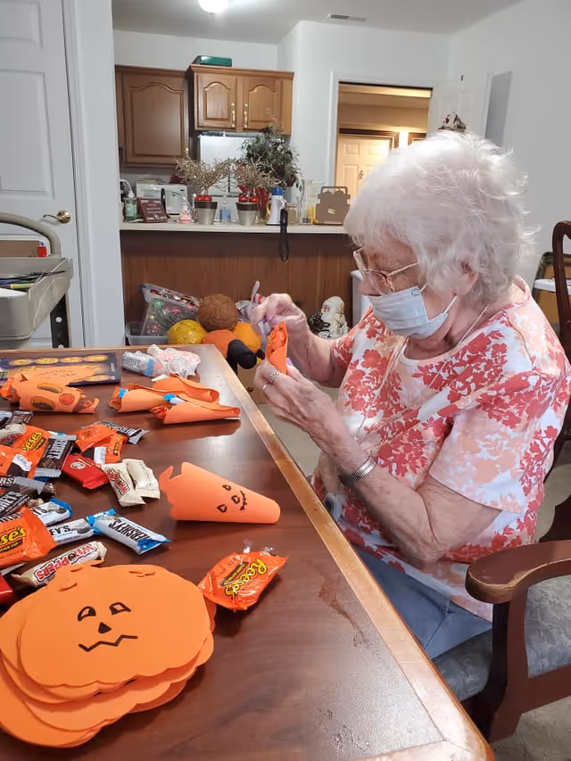 An elderly woman wearing a face mask and glasses is sitting at a wooden table in a room with a kitchen in the background. She is crafting orange paper cones decorated with jack-o'-lantern faces, surrounded by various Halloween-themed candy bars and paper pumpkin cutouts on the table.