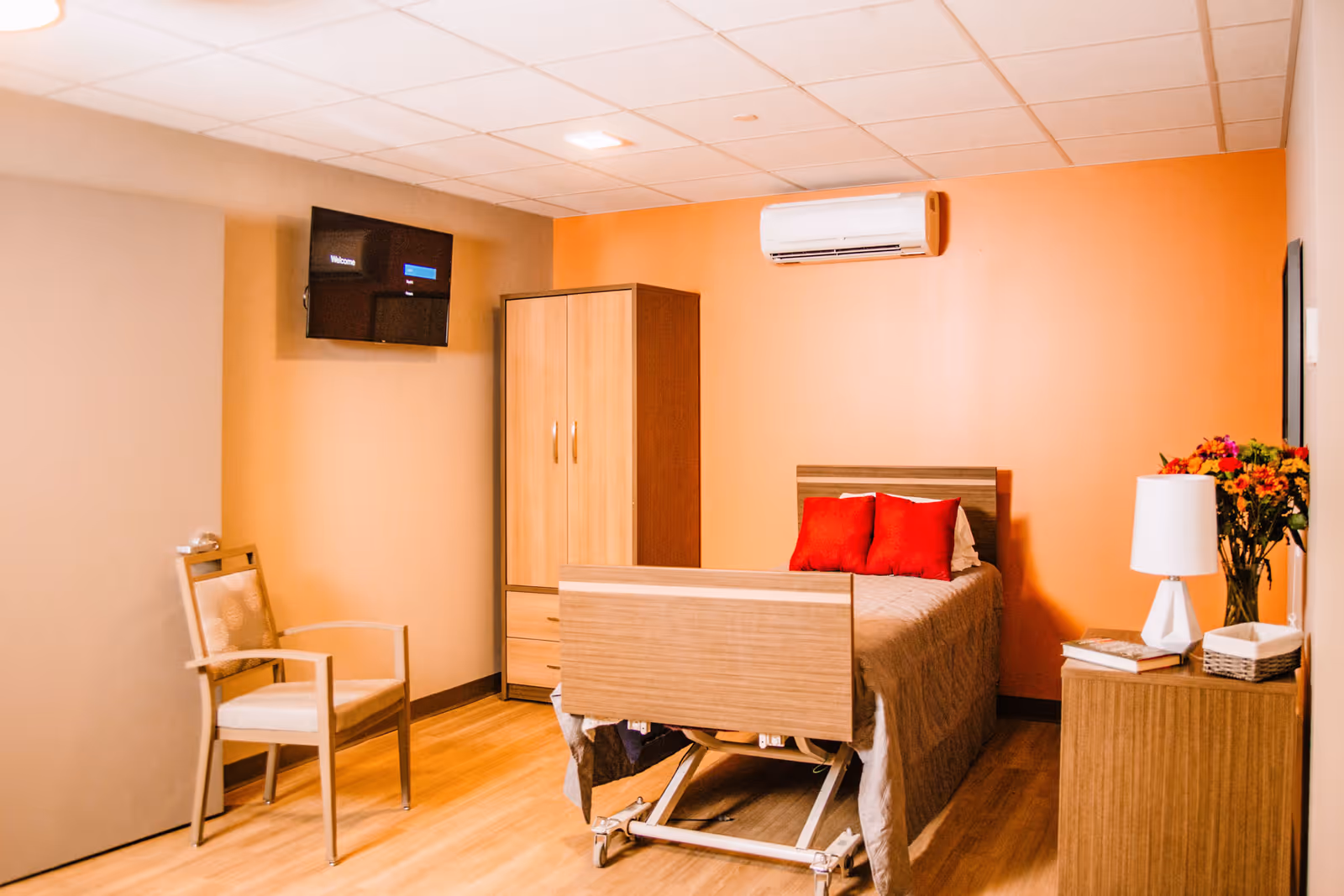 A cozy assisted living bedroom with a single bed featuring red pillows, a wooden wardrobe, a chair, a wall-mounted TV, an air conditioner, and a bedside table with a lamp and a vase of flowers.