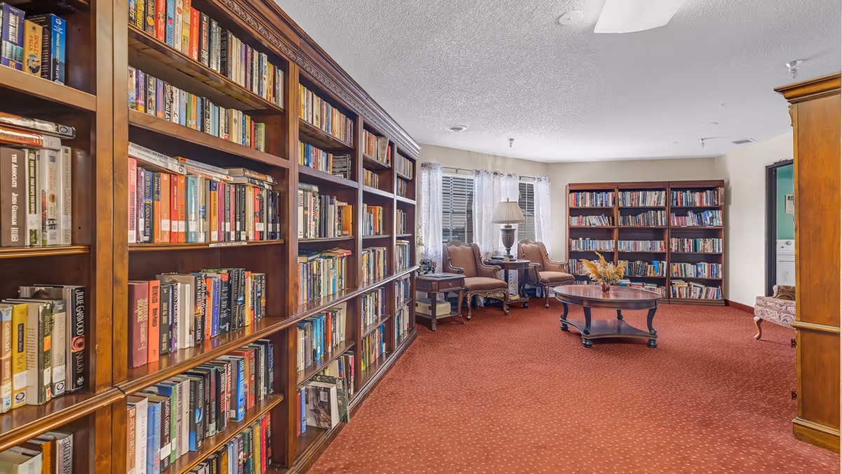 Wood-paneled library with rows of bookshelves, armchairs, and a round coffee table on red carpet.