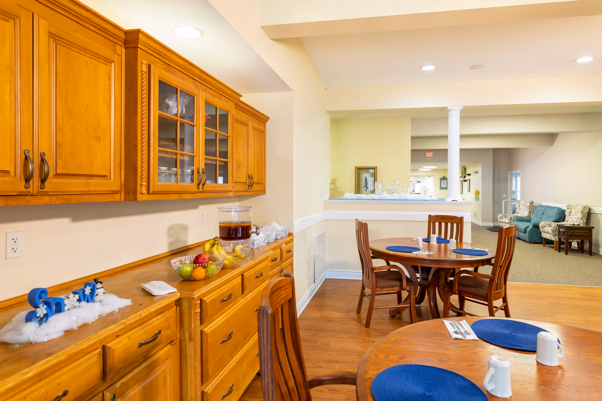 Interior view of a senior living facility dining area with wooden cabinets on the left side, a countertop with a beverage dispenser, fruit bowls, and decorative items. There are round wooden dining tables with blue placemats and chairs. In the background, there is a seating area with upholstered chairs and a sofa.