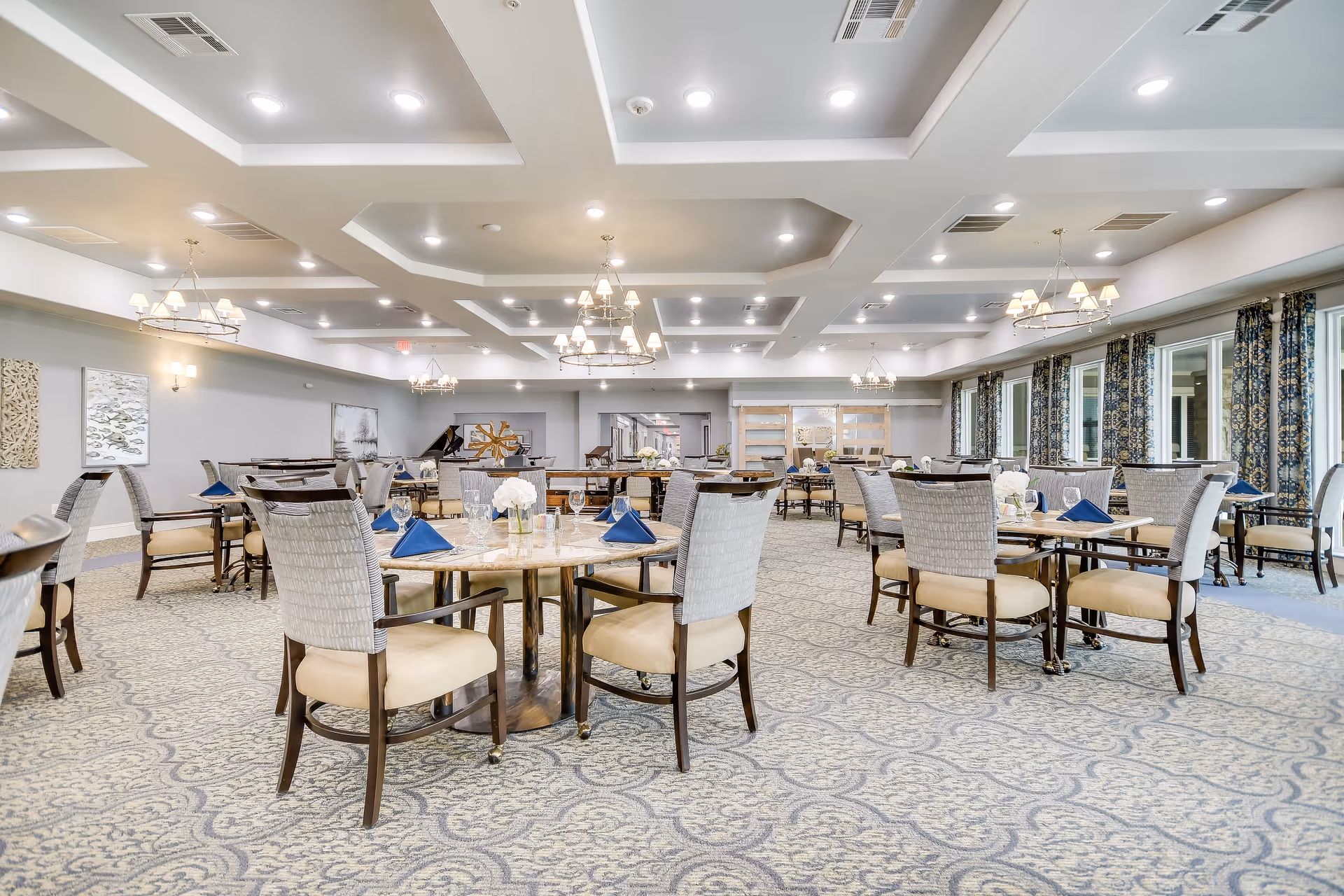 A spacious and elegant dining room in a senior living facility with multiple round tables set with blue napkins, glassware, and white floral centerpieces. The room features patterned carpet, large windows with floral curtains, and modern chandeliers hanging from a coffered ceiling.