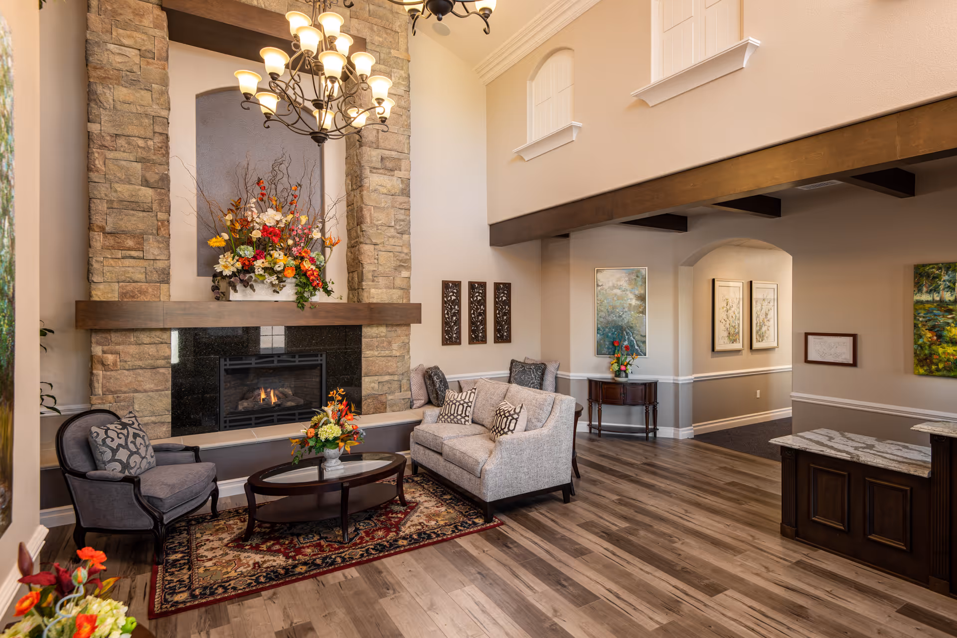 A cozy living room area in a senior living facility featuring a stone fireplace with a large floral arrangement on the mantel. The room has a beige sofa with patterned pillows, a gray armchair, a wooden coffee table with a flower vase, and a decorative rug. The walls are adorned with framed artwork, and there is a chandelier hanging from the high ceiling. The floor is wooden, and there is a reception desk visible on the right side.
