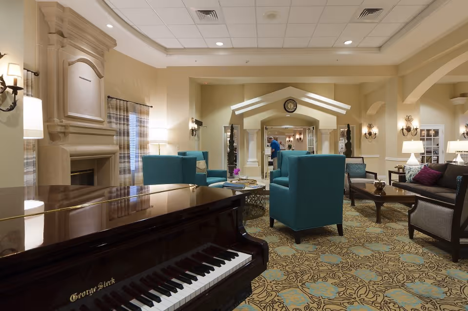 A spacious and elegant living room area in a senior living facility featuring a grand piano in the foreground, teal armchairs, a dark sofa with colorful cushions, a large fireplace, and warm lighting. The room has patterned carpet flooring and decorative wall sconces, with a person visible in the background near an archway.