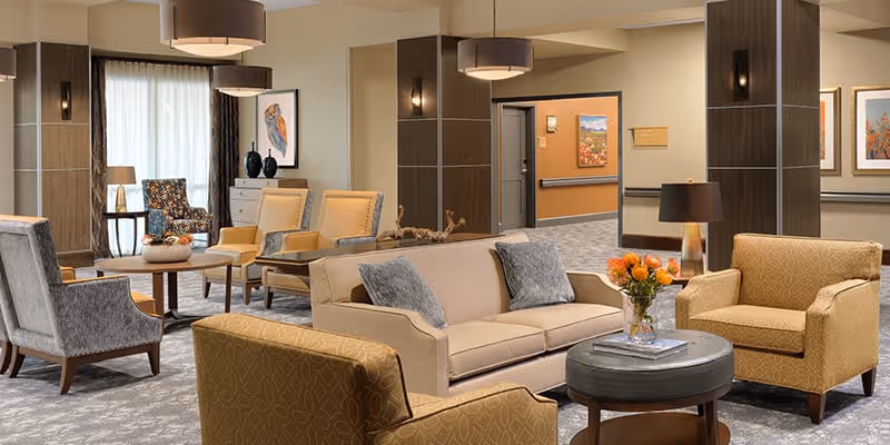 A spacious and well-lit senior living facility common area with comfortable seating including a beige sofa with gray pillows, yellow patterned armchairs, and gray chairs. There are round wooden tables with decorative items such as a vase with orange flowers and a lamp. The walls are decorated with framed artwork, and large windows with curtains allow natural light to enter. The area has modern light fixtures and a carpeted floor.