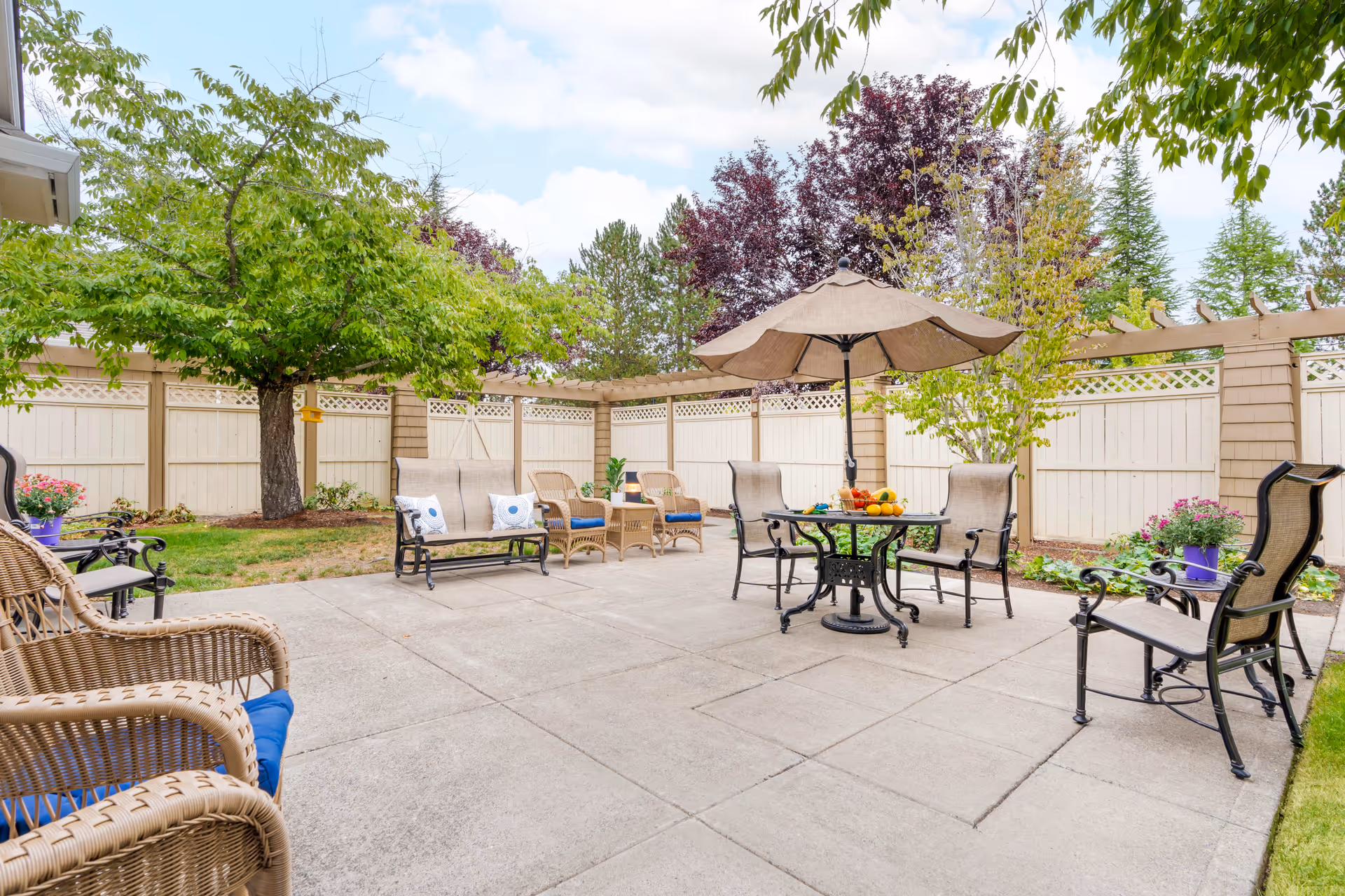 Outdoor patio area with various chairs and a table with an umbrella. The patio is surrounded by a white fence and trees with green and purple leaves. There are potted plants and flowers along the fence.