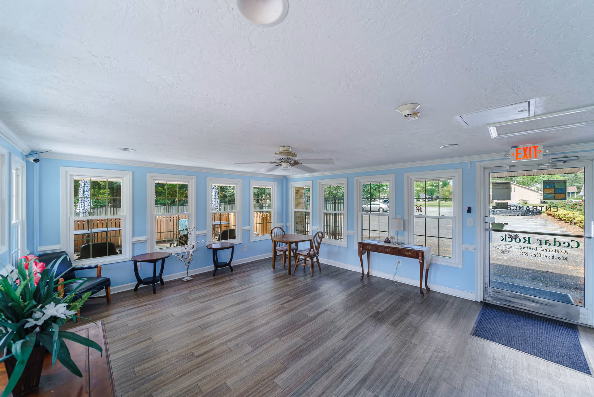 A bright and spacious room with light blue walls and multiple windows letting in natural light. The room features wooden flooring, a ceiling fan, a small table with two chairs, two small side tables, a wooden console table with a lamp, and a glass door with an exit sign above it. Outside the windows, a wooden fence and greenery are visible.