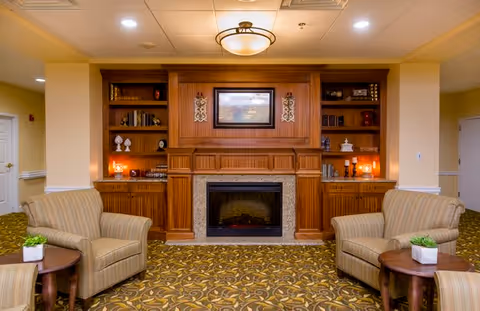 Cozy senior living common room with a central fireplace set in wooden built-ins, bookshelves, and two upholstered armchairs with side tables.