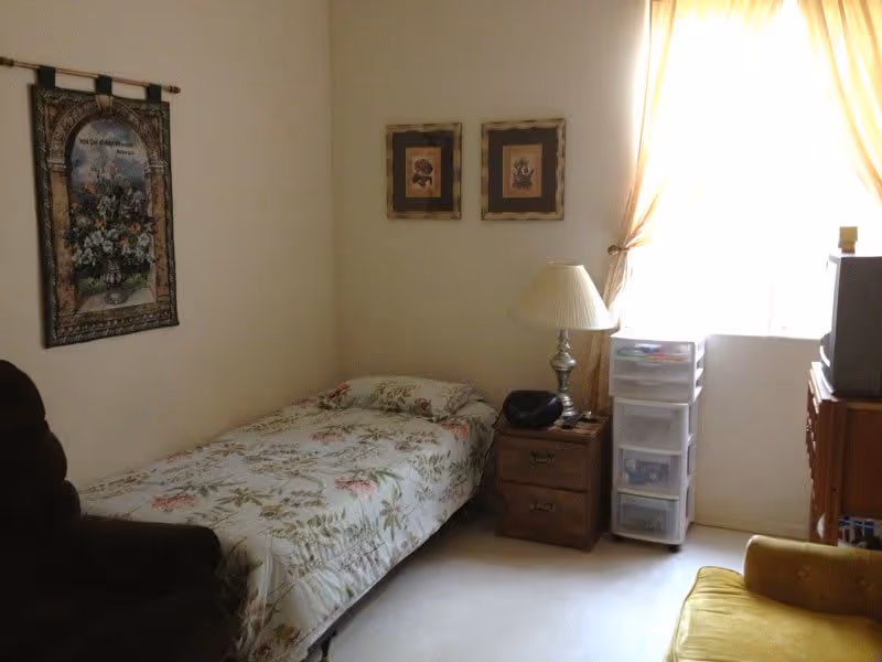 Small bedroom with a single bed covered in a floral bedspread, a nightstand with a lamp, plastic storage drawers and a TV by a curtained window.