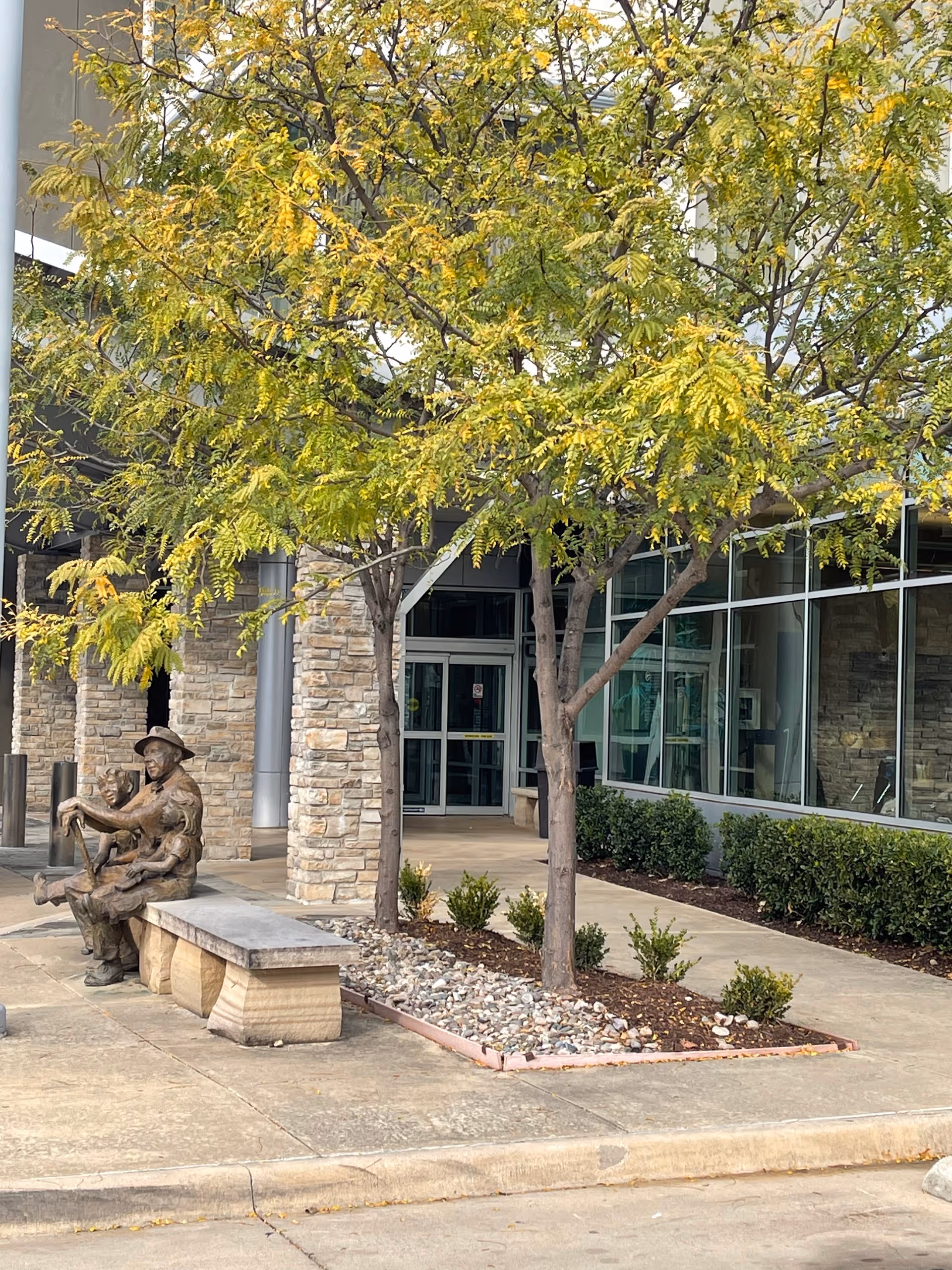 Entrance of a care facility showing stone pillars, glass-front facade, trees, and a bench with a bronze sculpture.