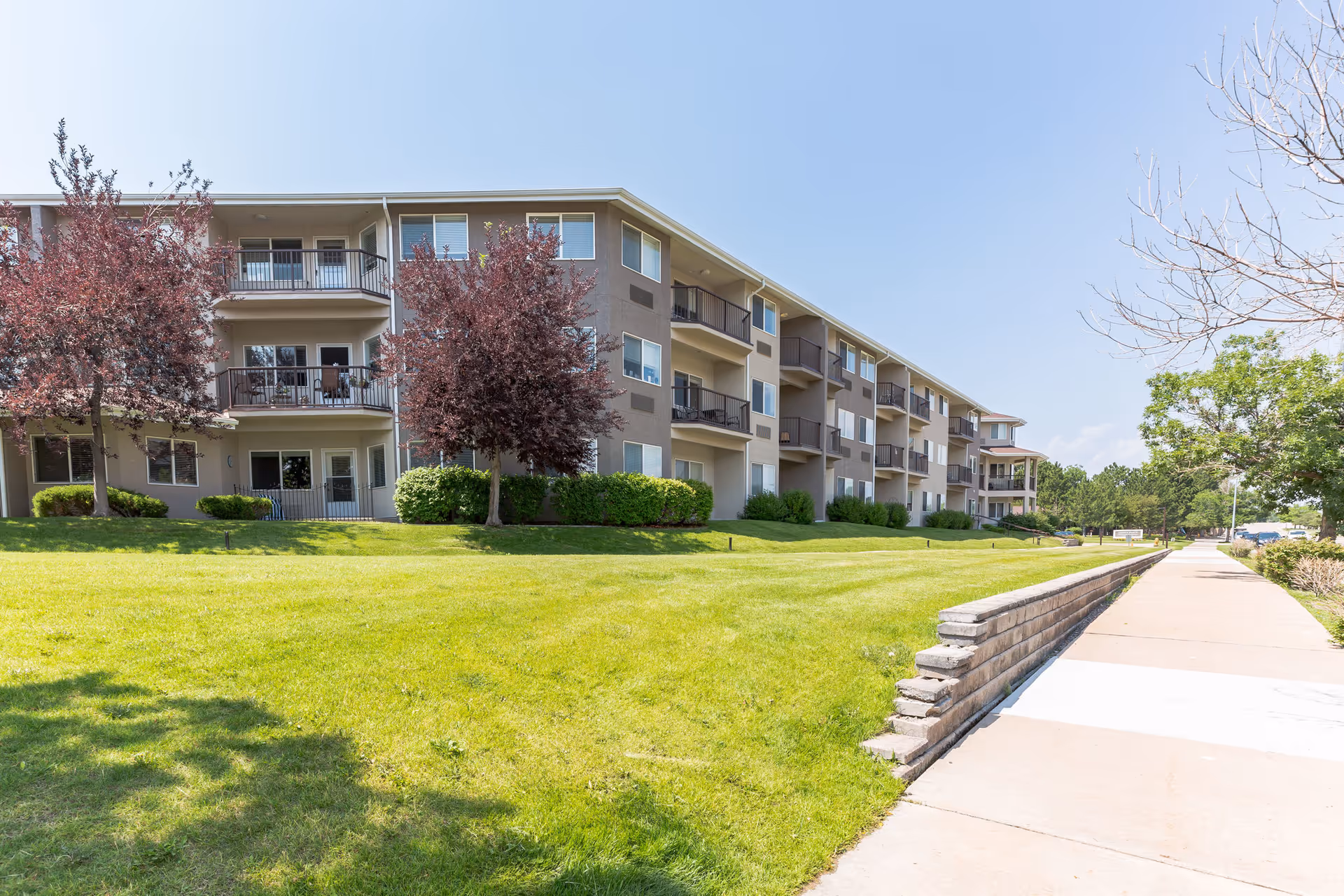 Exterior view of a multi-story residential building with balconies, surrounded by green grass, trees, and a sidewalk on a clear sunny day.