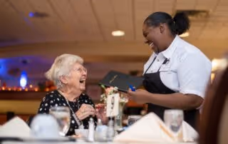An elderly woman sitting at a dining table in a restaurant or dining area, smiling and interacting with a waitress who is holding a notepad and pen. The table is set with glasses, plates, and napkins, and the background shows a warmly lit interior.