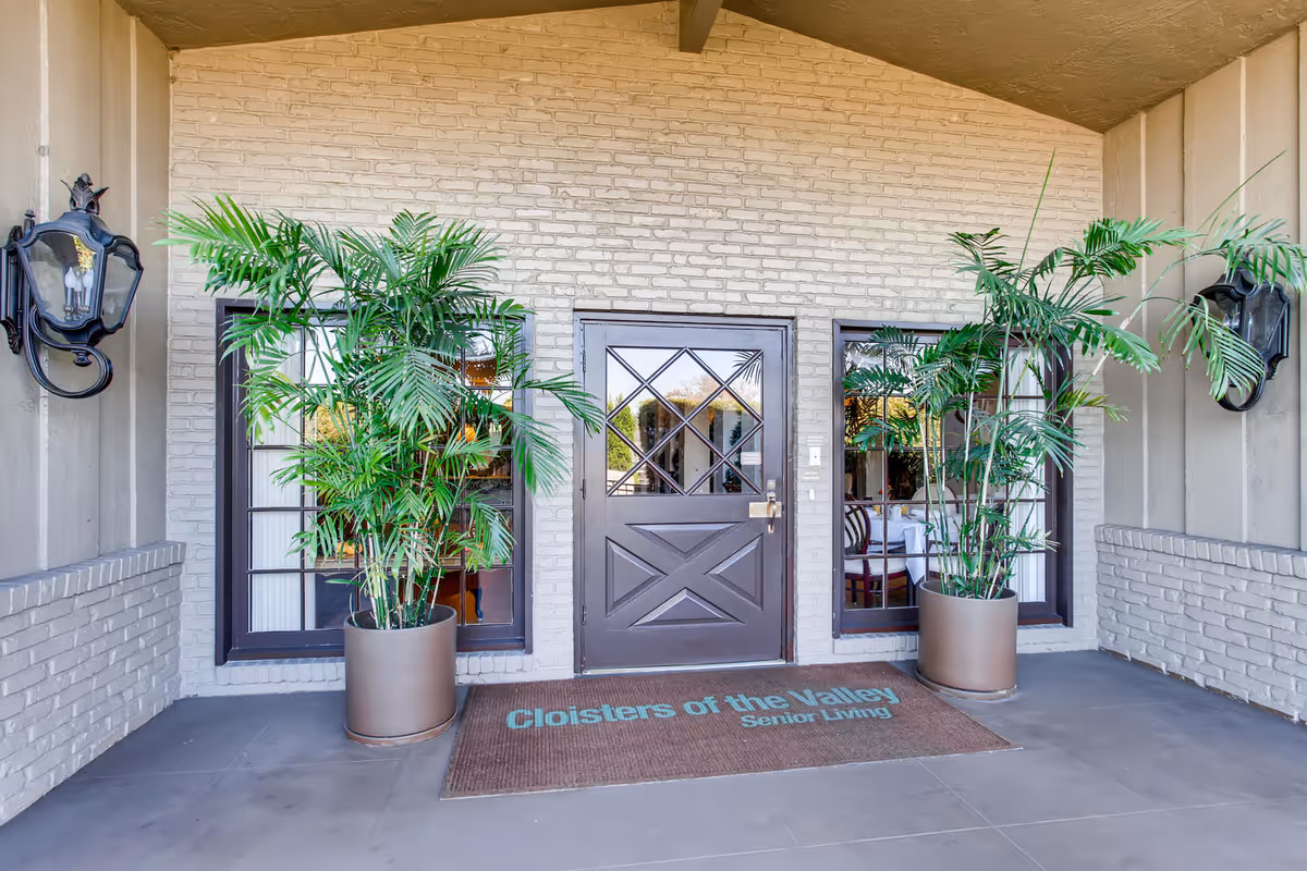 Entrance to Cloisters of the Valley senior living facility with a dark wooden door flanked by two windows and two large potted plants on either side. There are two black wall-mounted lanterns on each side of the entrance and a brown doormat with the facility name printed on it.