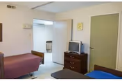 A simple shared patient bedroom with two beds, a dresser holding a small TV, a wall clock, and an open door leading to another room.