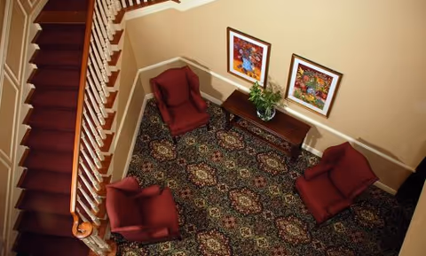 Overhead view of a stair landing with three red armchairs around a wooden console table, patterned carpet, framed artwork and a staircase.
