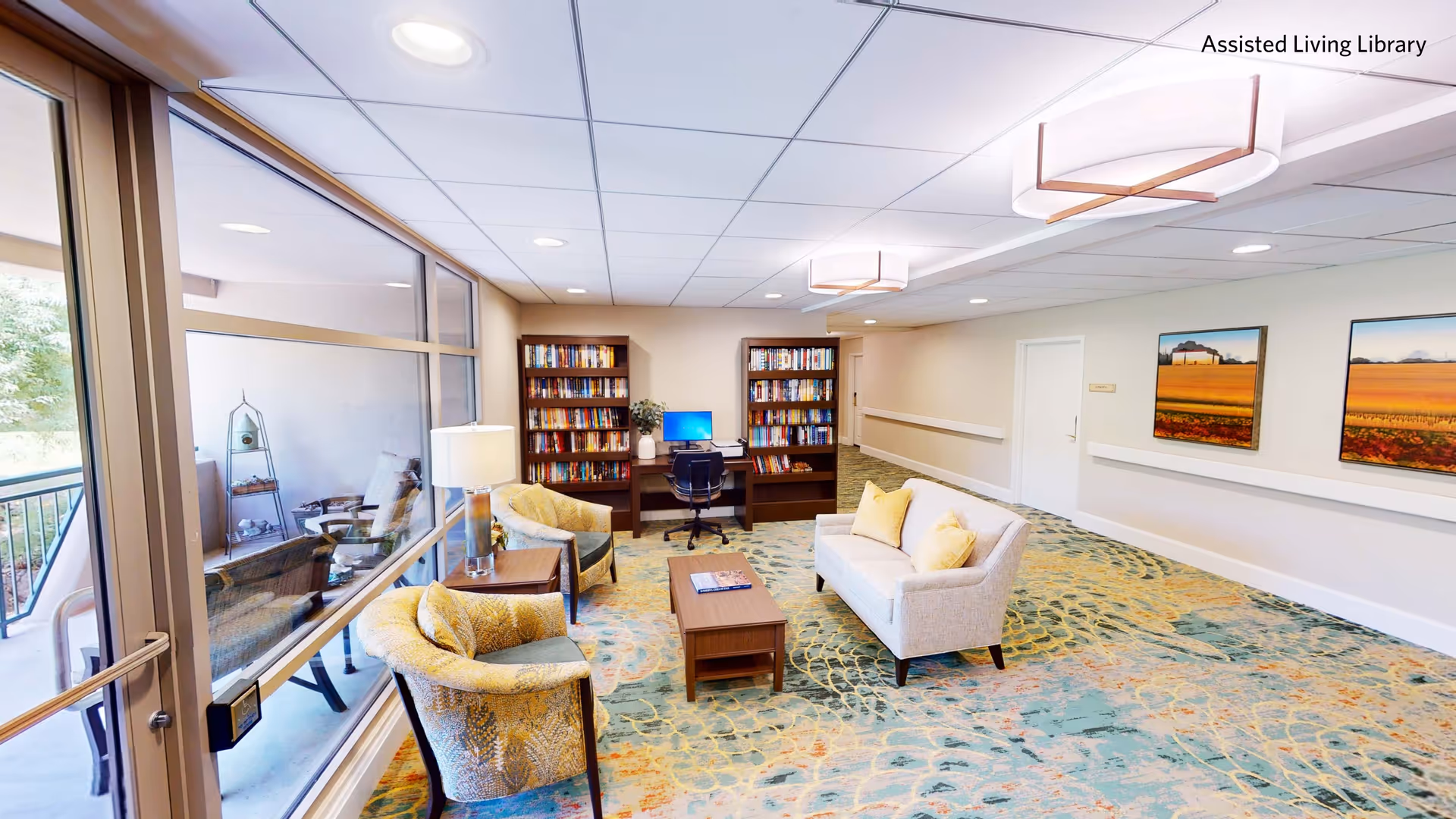 A bright assisted living library room with large windows on the left, two patterned armchairs, a small wooden coffee table, a light-colored sofa with yellow pillows, two tall bookshelves filled with books, and a desk with a computer and office chair. The room has colorful patterned carpet, ceiling lights, and two landscape paintings on the right wall.