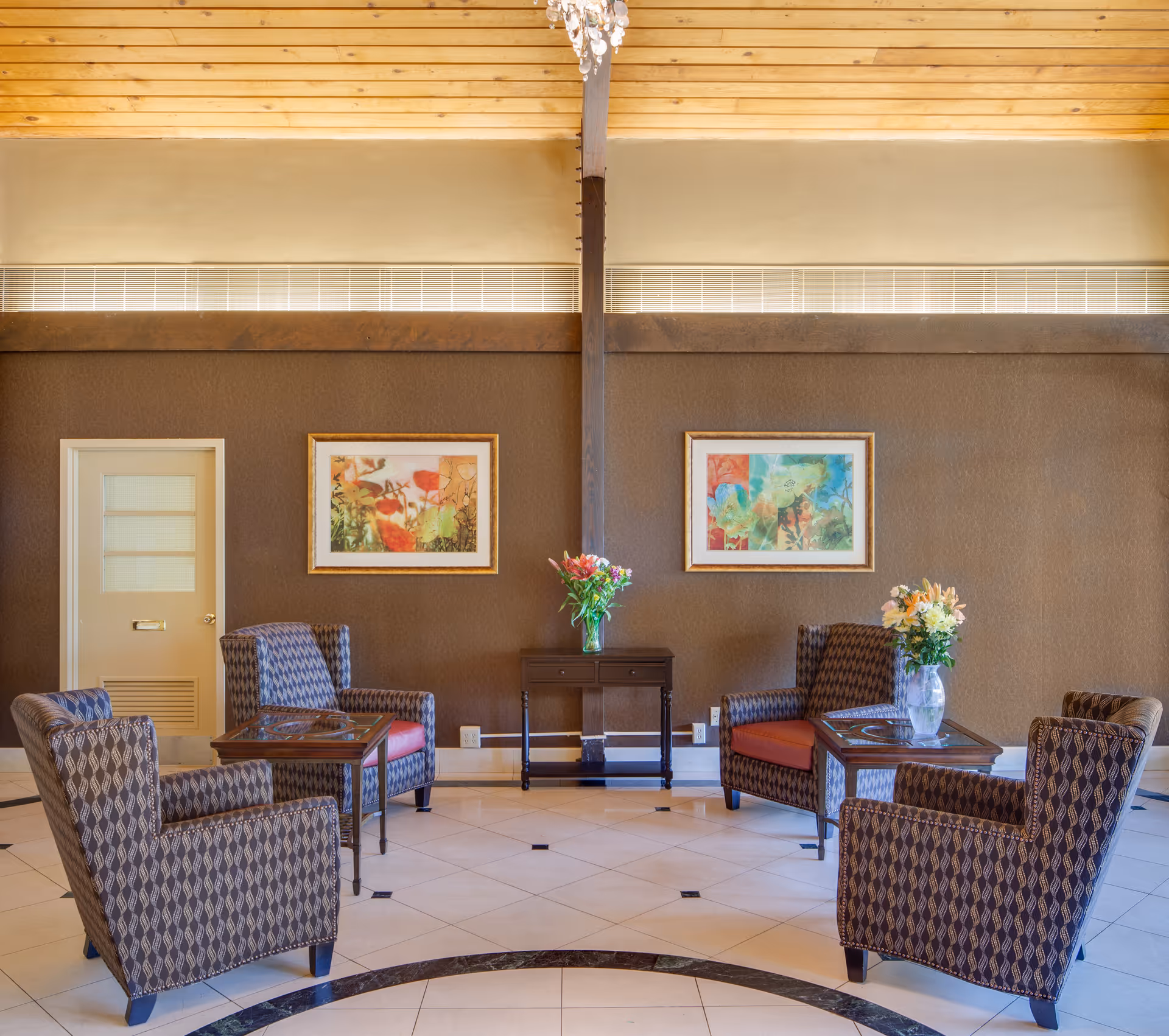A cozy sitting area with four patterned armchairs arranged around two small glass-top tables, each holding a vase with flowers. The room has a brown textured wall with two colorful framed paintings and a small wooden table with a flower vase in the center. The ceiling is wooden with a chandelier hanging down.