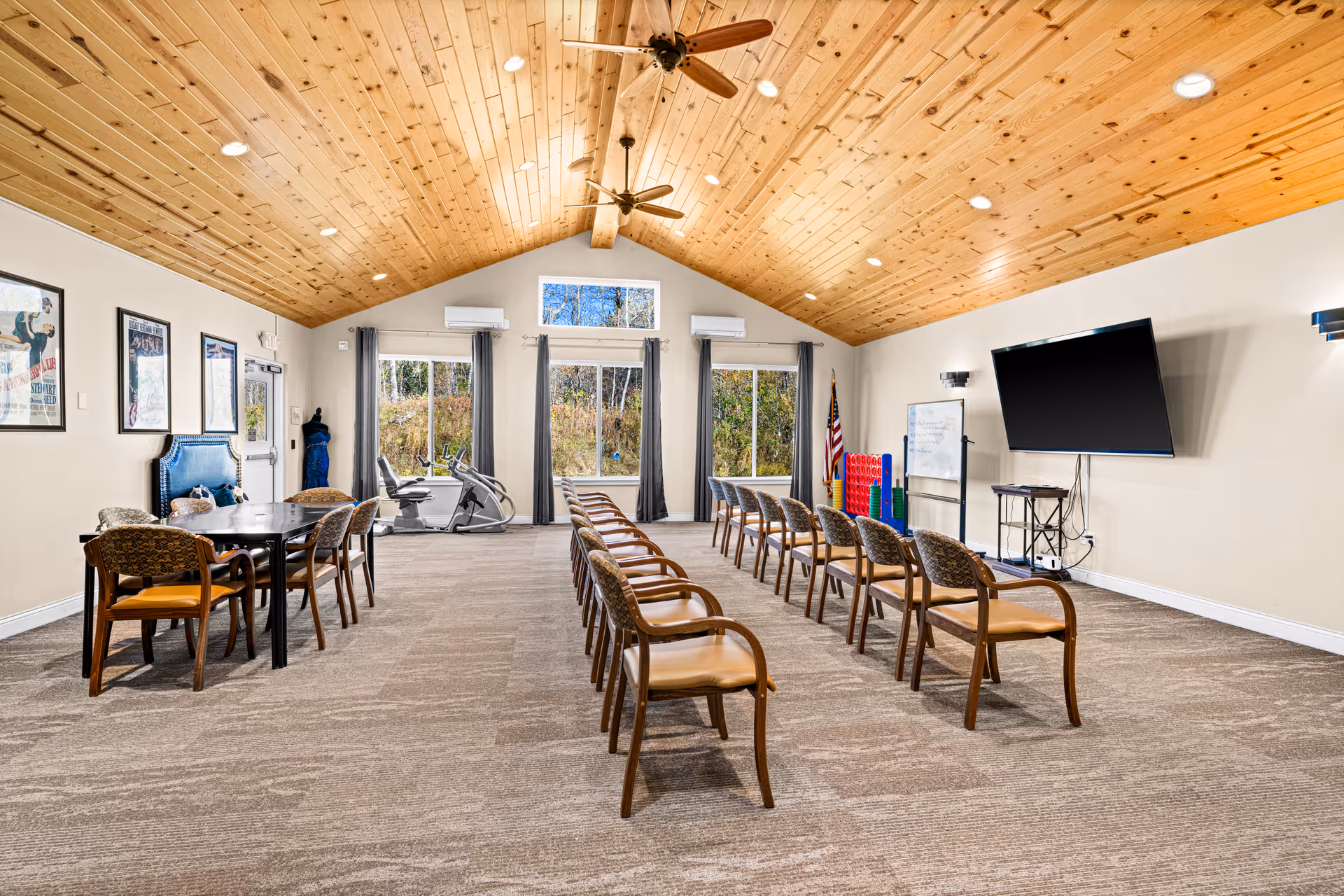 Spacious senior living activity room with rows of chairs facing a large wall-mounted TV, tables to the side, exercise equipment by the windows, and a wood-paneled vaulted ceiling.