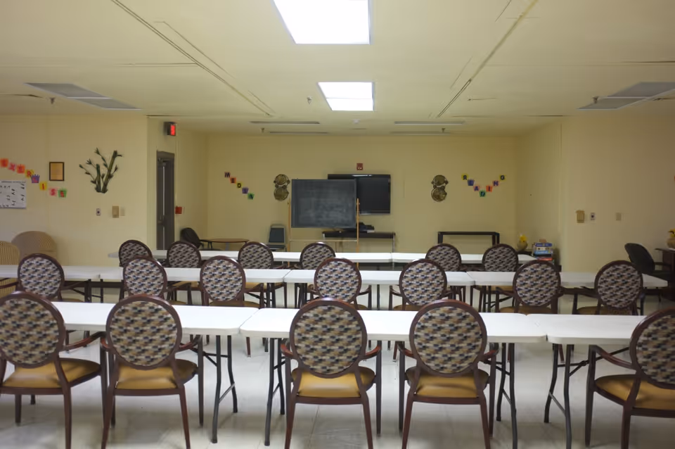 A room set up with multiple rows of white tables and patterned chairs facing a blackboard and a television mounted on the wall. The walls are light yellow with colorful letters and decorations. The ceiling has fluorescent lights.