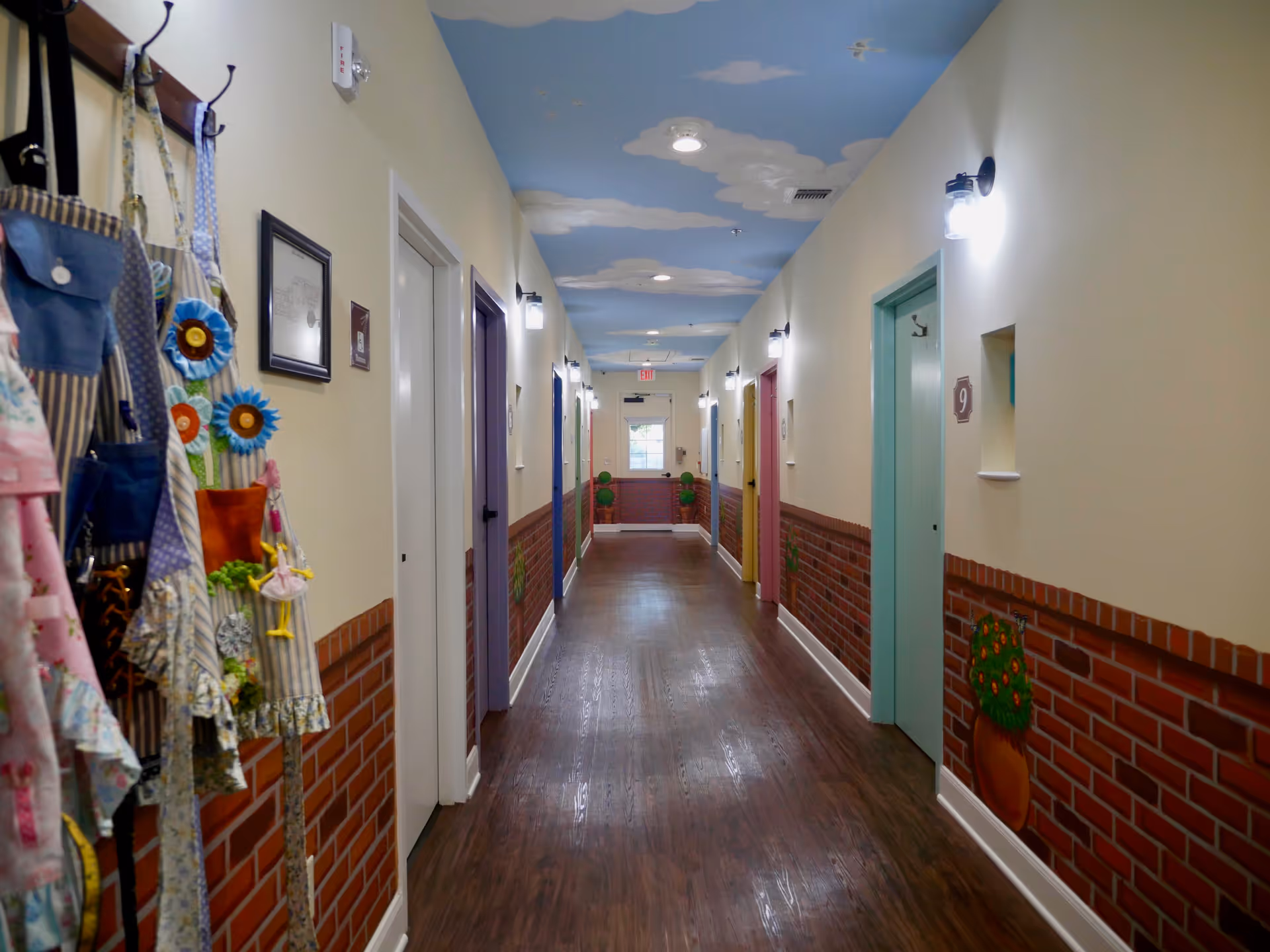 A brightly lit hallway in a senior living facility with multiple doors painted in pastel colors on both sides. The lower half of the walls is decorated with a brick pattern and flower pot murals. The ceiling is painted blue with white clouds, and aprons and bags hang on hooks on the left wall.