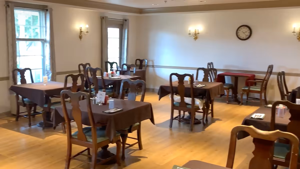 Dining room with several tables draped in brown tablecloths and wooden chairs in a senior living facility.
