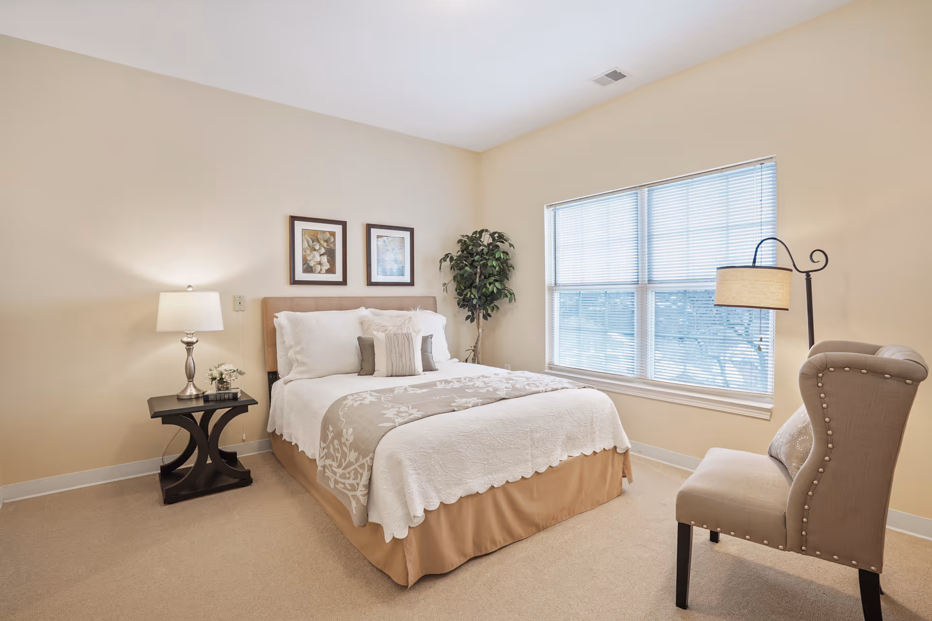 A bright bedroom with a neatly made bed featuring white and beige bedding, two framed pictures on the wall above the headboard, a small dark wooden nightstand with a lamp and a small plant, a large window with blinds, a tall floor lamp, a beige upholstered chair with nailhead trim, and a potted plant in the corner.