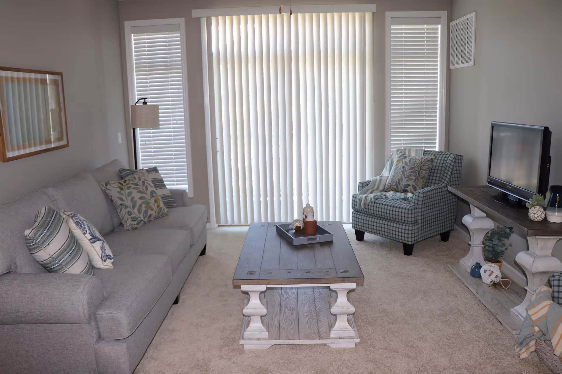 A bright living room with a gray sofa, patterned armchair, wooden coffee table, TV on a console, and vertical blinds over sliding glass doors.
